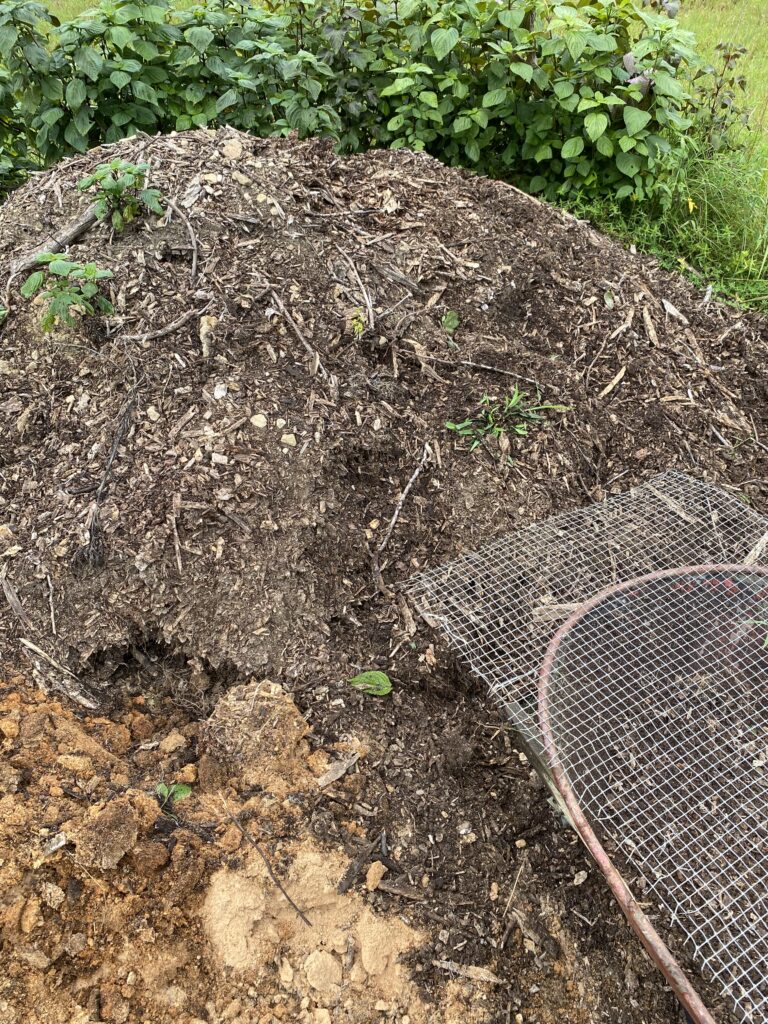 Large mound of dark, decomposing wood chips with a wire mesh sifting screen leaning against it