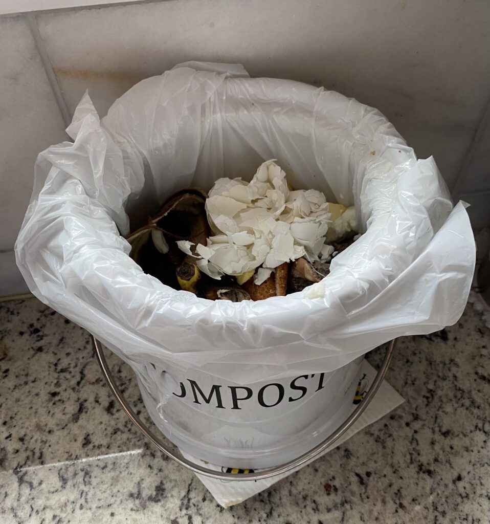 Countertop compost pail lined with a white bag, full of eggshells and banana peels